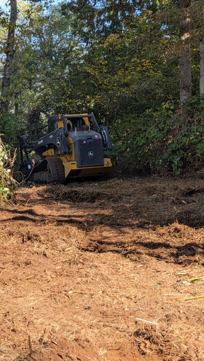 John Deere track loader mulching brush at the edge of a wooded lot, with a fresh layer of wood chips on the ground in front of it